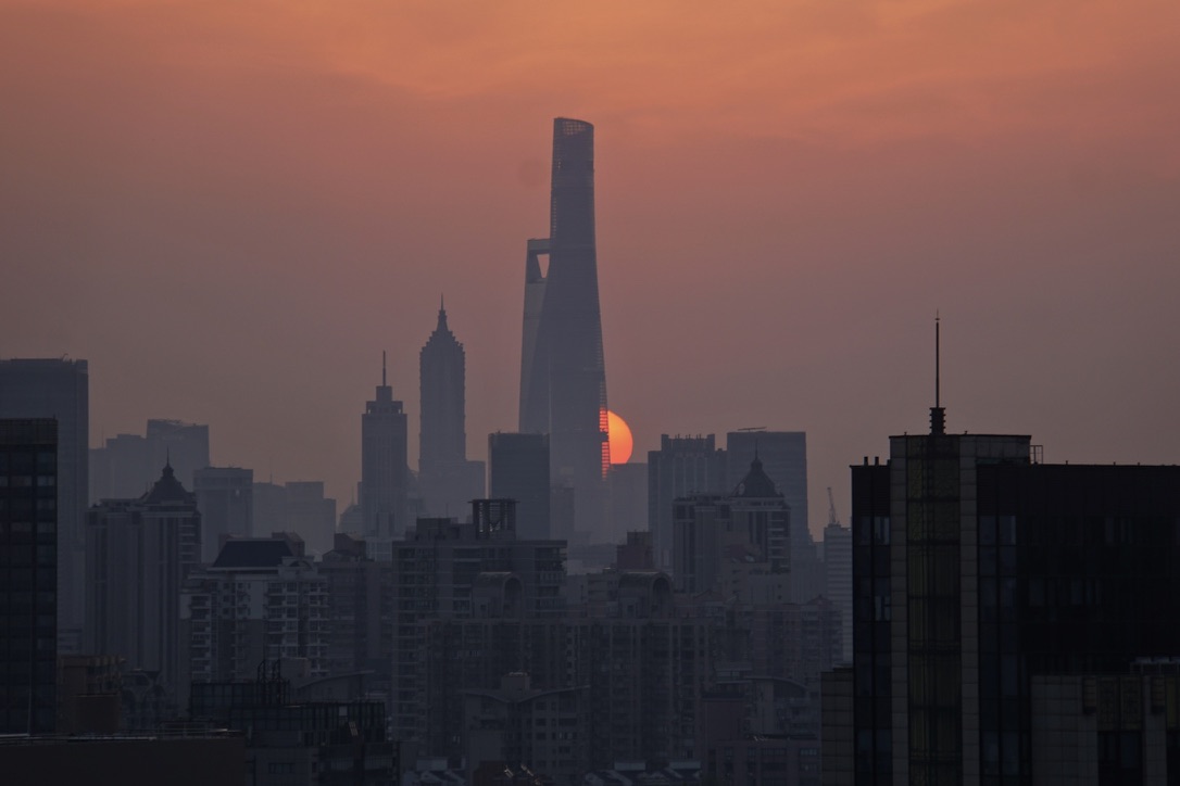 The sun setting orange behind the Shanghai Tower in a hazy city skyline at dusk