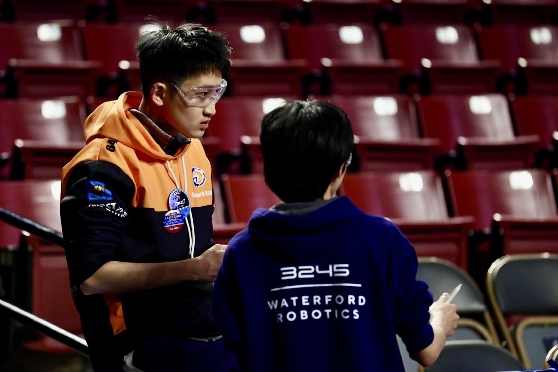 Jackson in an orange team shirt and safety glasses talking strategy with a teammate at a FIRST Robotics competition